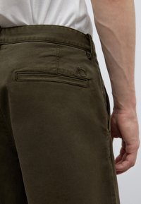 Right side of person wearing olive green pants with back slit pocket and visible white shirt tucked in, arm relaxed by side.