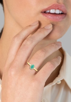 Goldring mit einem ovalen grünen Stein, umgeben von kleinen klaren Steinen, getragen an einer Hand, die das Kinn berührt, mit glatter Hautstruktur.