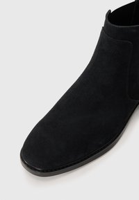 Bota de tobillo de ante negro con punta redondeada, textura suave y suela negra. Detalles de costura mínimos cerca de la abertura.