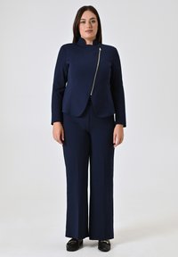 Marineblauwe, getailleerde jas met een hoge kraag en een schuine rits, gecombineerd met rechtvallende marineblauwe broek. Glad stofoppervlak. Zwarte schoenen.