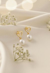 Guldörhängen i hjärtform med en pärldroppe och en liten kristalldetalj. Placeras mot en mjuk, krämfärgad tygbakgrund.