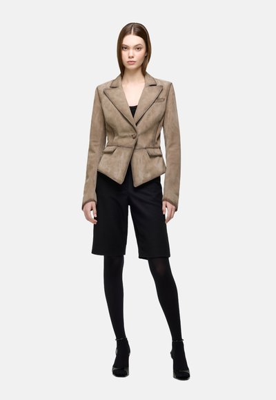 Beige suede blazer with a fitted design, notch lapels, and two front pockets. Styled over black shorts and opaque tights, paired with heels.