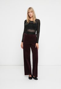 Femeie îmbrăcată cu un top lung cu mâneci transparente de culoare neagră, pantaloni larghi de un burgundy închis cu talie înaltă și balerini negri, stând pe un fundal alb.