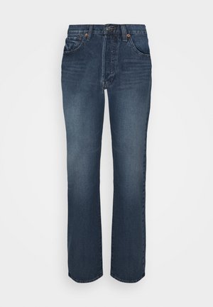 Hvid kortærmet t-shirt med rød logo, blå denim jeans, grønne og hvide sneakers og en gul baseballcap holdt i hånden.