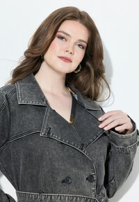 Šedý denimový trenčkot s dvojitým zapínáním, velkým límcem, černými knoflíky a jemnými švy. Model si sahá na límec.