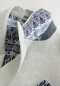 Blazer gris clair texturé avec un col intérieur à motif blanc, bleu et noir, accompagné d'un mouchoir de poche assorti.