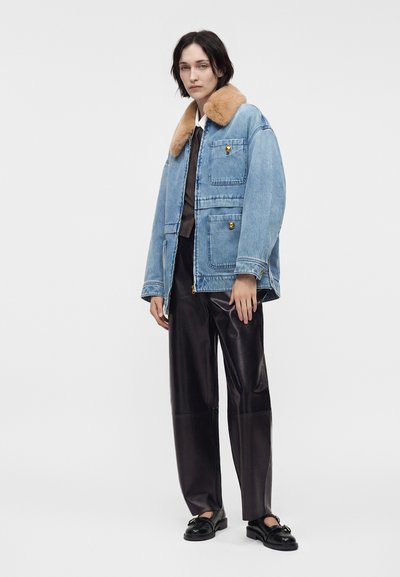 Personne portant une veste en jean bleue avec un col en fourrure beige, un pantalon en cuir noir et des mocassins noirs, debout devant un fond gris clair.