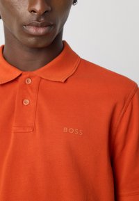 Nærbilde av en person som har på seg en oransje polo-skjorte med to knapper og en liten "BOSS" logo på brystet.