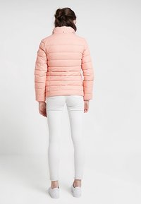 Brzoskwiniowa kurtka puchowa z poziomym pikowaniem, wysokim kołnierzem i zatrzaskami. Nosi się ją na biały obcisły dżins i białe trampki.