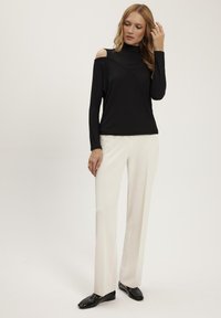Czarna bluzka z długim rękawem i wycięciami na ramionach, wysoki kołnierz i krzyżowe detale, w połączeniu z białymi spodniami prostymi i czarnymi loafersami.