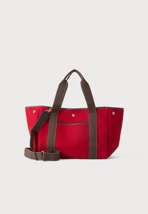 Rød canvas-tote taske med mørkebrune håndtag og justerbar skulderrem, der hviler på en lys baggrund.