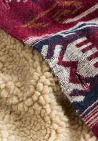 Fleece tæppe med en blød beige underside og et mønstret uldtop i burgunder, marineblå og creme, der har geometriske designs og tekstureret stof.