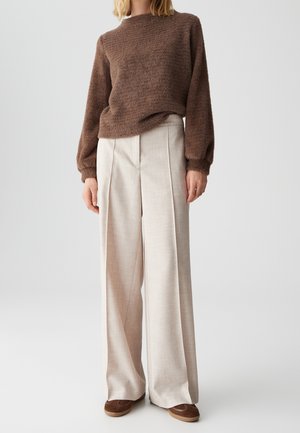 Personne portant un pull marron duveteux et un pantalon beige large plissé avec des chaussures marron sur un fond uni.