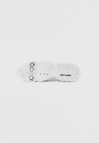 Bílé sportovní boty s texturovanou gumovou podrážkou, obsahující výrazný detail loga a vzorované akcenty na spodní straně.