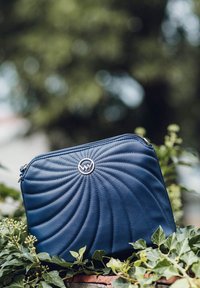 Námořnická modrá kožená taška se spirálovým prošíváním a stříbrným zipem, s kulatým logem na přední straně. Texturovaný povrch, obdélníkový tvar.