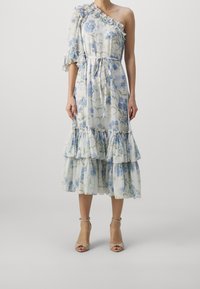 Robe florale à une épaule avec une manche à volants, présentant un fond blanc, des fleurs bleues et un ourlet à volants en trois niveaux. Détail à la taille nouée.