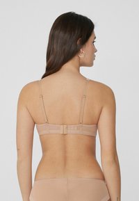 Hnědá podprsenka v nahé barvě s nastavitelnými ramínky, zapínáním na háček a oko, a hladkým, texturovaným materiálem. Záda mají jednoduchý design s minimálními detaily.