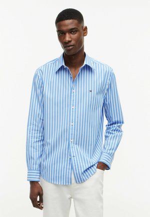 Mann trägt blau-weiß vertikal gestreiftes Hemd mit Knöpfen und weiße Hose, steht vor einem schlichten weißen Hintergrund.