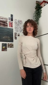 Top beige a maniche lunghe con un design della vita annodato e tessuto testurizzato, abbinato a jeans scuri. Lo sfondo include opere d'arte murale e piante.
