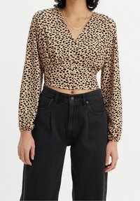 Korte blouse van beige satin met zwarte luipaardvlekken, v-hals, lange puffmouwen en knopen aan de voorkant; gecombineerd met zwarte high-waisted jeans.