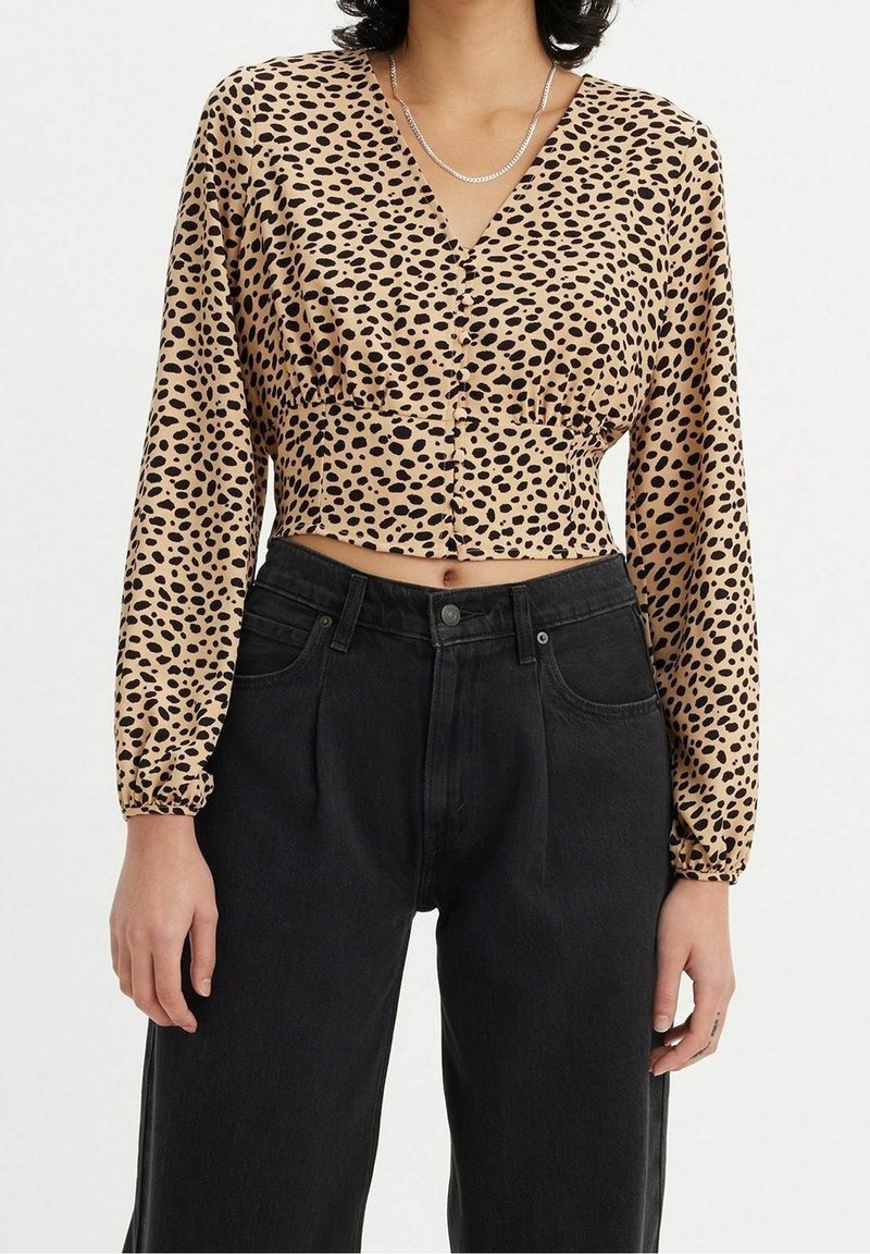 Korte blouse van beige satin met zwarte luipaardvlekken, v-hals, lange puffmouwen en knopen aan de voorkant; gecombineerd met zwarte high-waisted jeans.