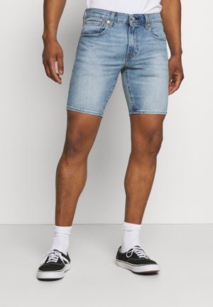Shorts en denim bleu clair avec une coupe slim, taille mi-haute, cinq poches et des zones légèrement décolorées. Associés à des baskets noires et des chaussettes blanches.