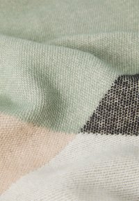 Nærbillede af strikket stof med bløde grønne, beige, sorte og hvide farveblokke, der viser tekstur og vævdetaljer.