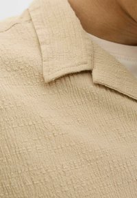 Tela texturizada beige con un patrón arrugado; presenta un cuello puntiagudo y costuras visibles. Primer plano del detalle de la superficie de la prenda.