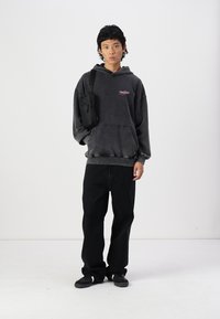 Prekračovaný čierny hoodie s logom, vyblednutou textúrou, kengurskou kapsou; v páre s voľnými čiernymi džínsami a čiernymi teniskami. Taška na ramene.