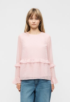 Jeune femme aux cheveux blonds raides, portant un chemisier transparent à manches longues rose clair avec des détails en volants et un jean bleu, debout devant un fond blanc.