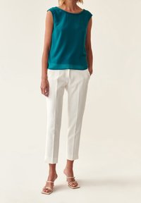Top sin mangas en color azul verde con escote en forma de ondas, combinado con pantalones blancos ajustados y tacones beige con tiras. Tela suave y silueta ajustada.