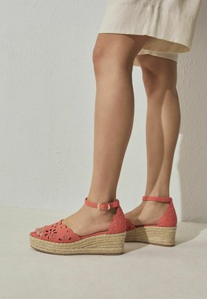 Femme portant des sandales compensées corail avec des brides à la cheville et des découpes florales, associées à une jupe beige devant un mur clair uni.