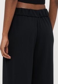 Schwarze, hoch taillierte Hose mit einem gerafften Bund. Hergestellt aus einem glatten, dehnbaren Material. Seitennaht sichtbar, die eine lockere Passform zeigt.