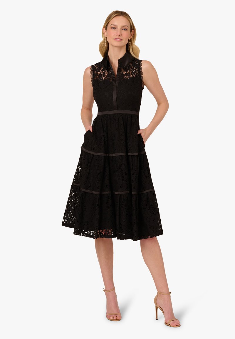Abito in pizzo nero con scollo a collare, design senza maniche, gonna a strati e due tasche laterali. Accentuato da dettagli in nastro di raso.