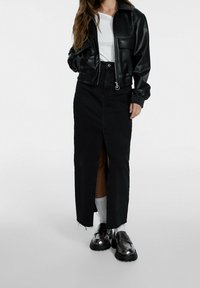 Veste cropped en faux cuir noire avec fermeture éclair, jupe maxi en denim noir avec fente avant, et mocassins à plateforme noirs brillants avec des chaussettes blanches.