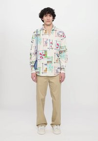 Button-up shirt in light cream cotton with colorful beach-themed patterns. Paired with beige trousers and white sneakers.