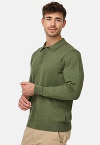 Polo de manga larga verde hecho de una tela suave y texturizada. Cuenta con un cuello, una tapeta de tres botones y puños acanalados para un ajuste ceñido.