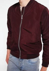 Bordeauxfarbener Bomberjacke mit Reißverschluss auf der Vorderseite, geripptem Kragen und Bündchen sowie seitlichen Taschen; aus glattem, leichtem Stoff, getragen über blauen Jeans.
