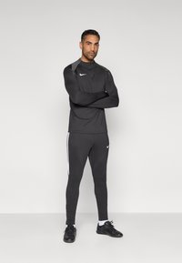 Μαύρο αθλητικό μακρυμάνικο τοπ και εφαρμοστά παντελόνια με λευκές πλευρικές ρίγες. Διαθέτουν λογότυπο Nike και στα δύο ρούχα. Λεία υφή, αθλητικός σχεδιασμός.