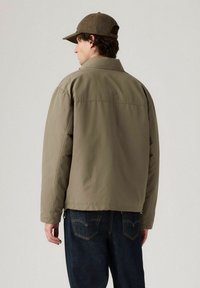 Man i brunt cap, olivgrön jacka och mörkblå jeans stående med ryggen mot kameran framför en neutral bakgrund.