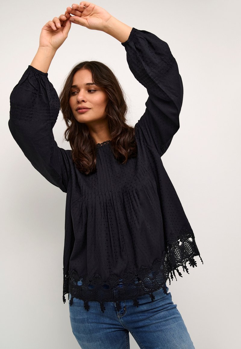 Melna blūze ar garām piedurknēm, teksturēts audums, plāksnes un mežģīņu apdare apakšmalā. Nēsāta ar zilām džinsiem.