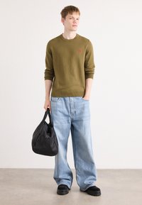Pull vert olive surdimensionné, jeans baggy bleu clair, et chaussures noires. Accessoire : sac en tissu noir avec poignées. Design minimaliste.
