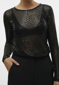 Svart genomskinlig långärmad topp med ett mönster av diamantpaljetter, bärs över en svart bralette, ihop med svarta högmidjade byxor och en hand i fickan.