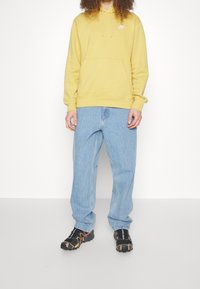 Sudadera amarilla con capucha y bolsillo frontal, combinada con jeans de mezclilla azul claro y zapatillas deportivas negras con detalles en naranja.