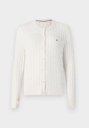 Fehér kötött kardigán hosszú ujjakkal, kerek nyakkivágással és gombos záródással. Bordás mandzsettákkal és szegéllyel rendelkezik, valamint egy kis logó található a mellen.