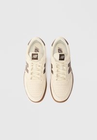 Kremowe sneakersy z brązowymi akcentami, wykonane z zamszu i siateczki, z perforowaną częścią nosową, płaskimi sznurowadłami oraz logo "NB" na języku.