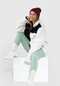 Giacca in pile bianca e nera, leggins verde menta, calzini gialli con dettagli neri e sneaker bianche. Modello seduto su un blocco bianco.