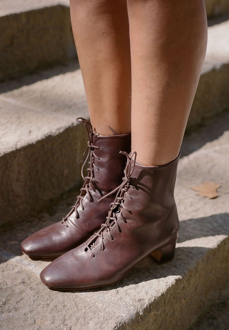 Brązowe skórzane botki za kostkę z noskiem w szpic, zawiązywane na sznurówki, z niewielkim obcasem typu blok. Faktura gładka z lekko postarzaną powierzchnią.