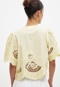 Svetlo rumena majica z zapletenimi čipkastimi izrezi z cvetličnimi vzorci. Ohlapen kroj z napihnjenimi rokavi in drljanimi robovi.