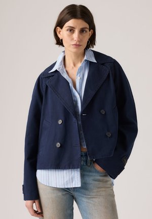 Femme aux cheveux courts et foncés portant une veste bleu marine à double boutonnage sur une chemise à rayures bleu clair avec boutons et un jean bleu délavé, debout de manière détendue.